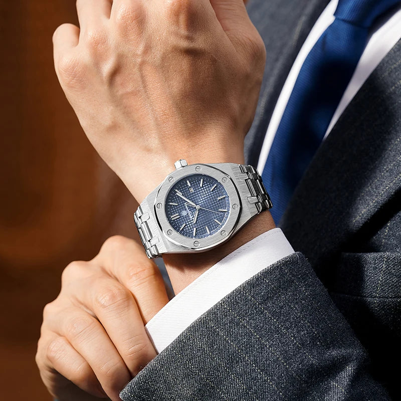 A close-up of a man wearing a silver square quartz watch on his wrist, paired with a dark suit jacket and blue tie.