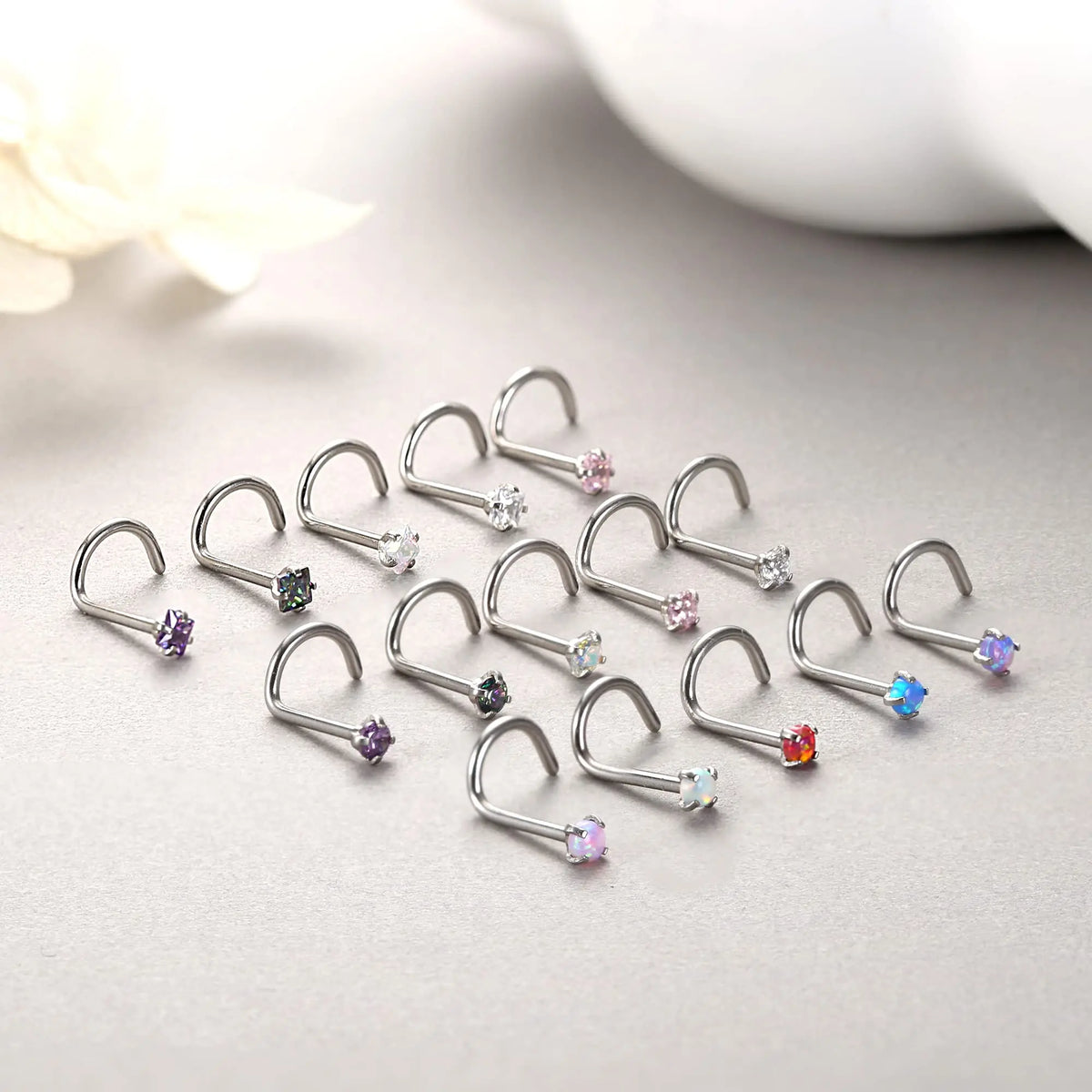 A collection of surgical stainless steel nose piercing studs with various colored gemstones arranged on a light gray surface.