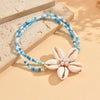 A beaded bracelet featuring white and blue cowrie shells with a floral charm, displayed on a light surface.