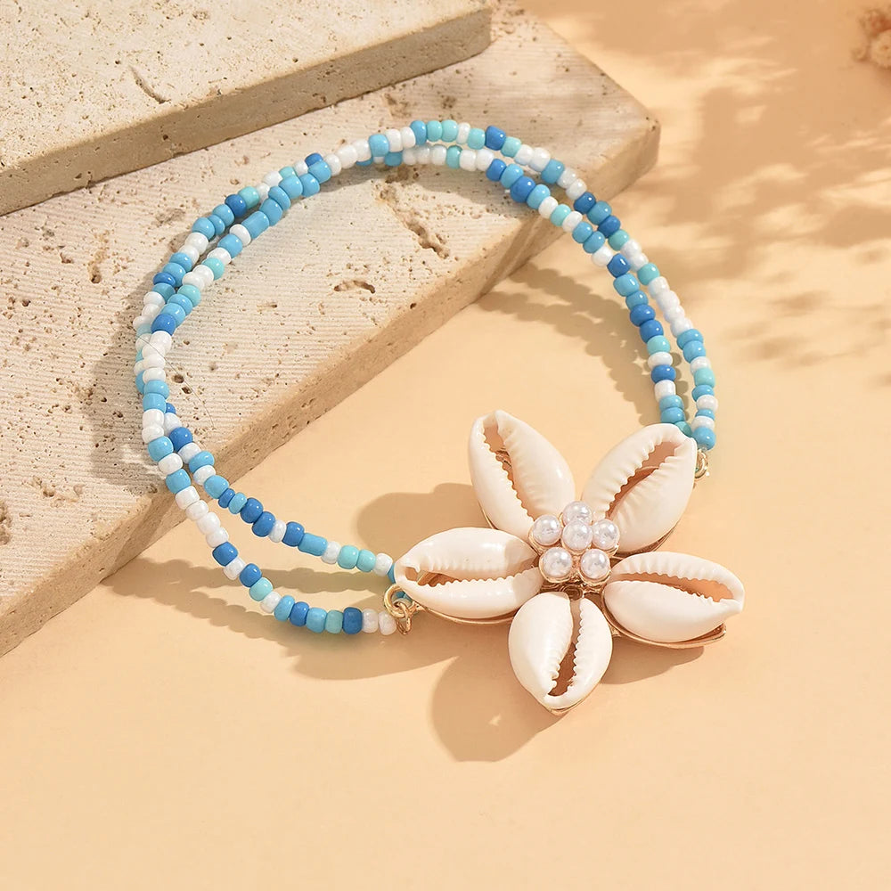A beaded bracelet featuring white and blue cowrie shells with a floral charm, displayed on a light surface.