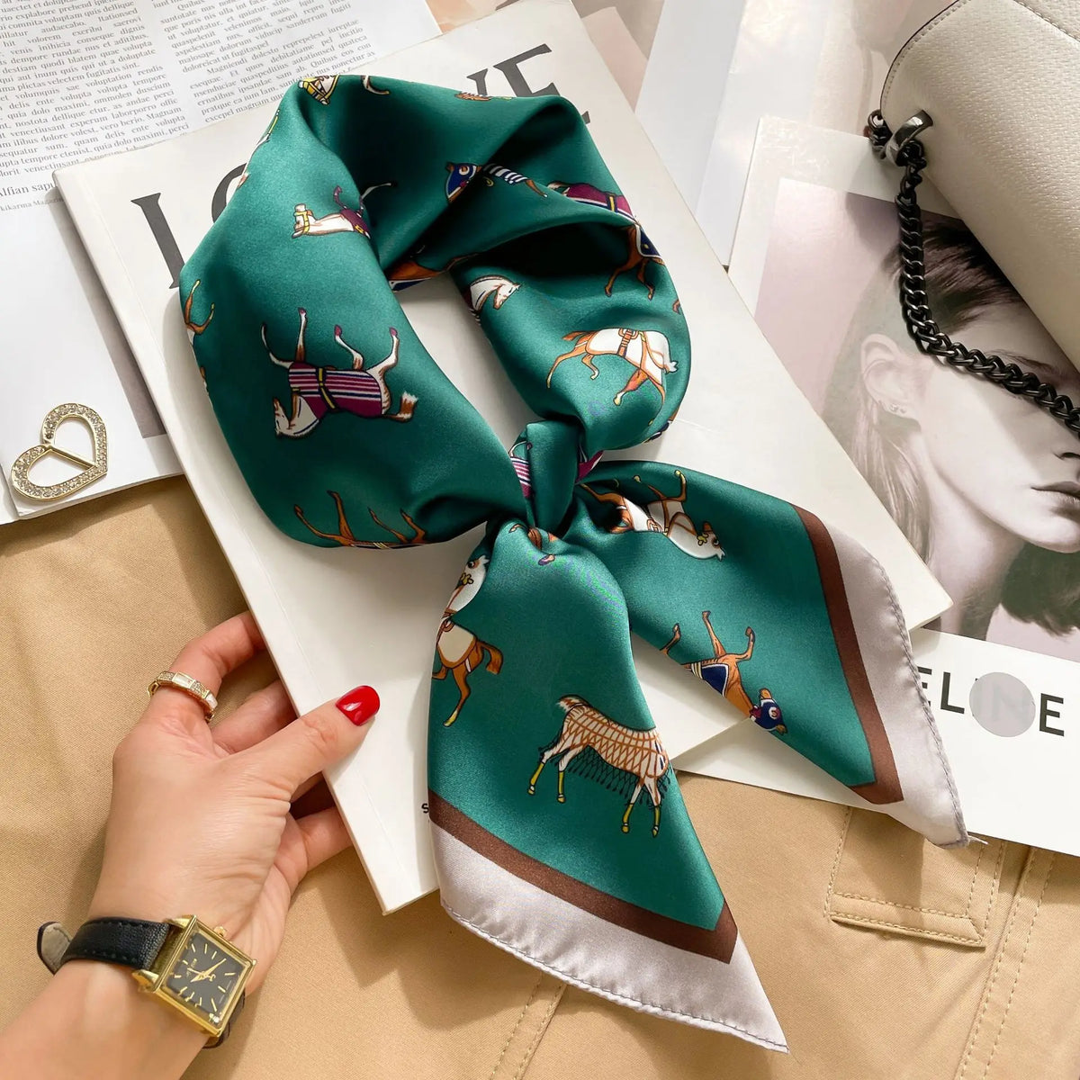A teal silk scarf tied in a bow is displayed on a white card next to a gold watch and a light beige jacket.