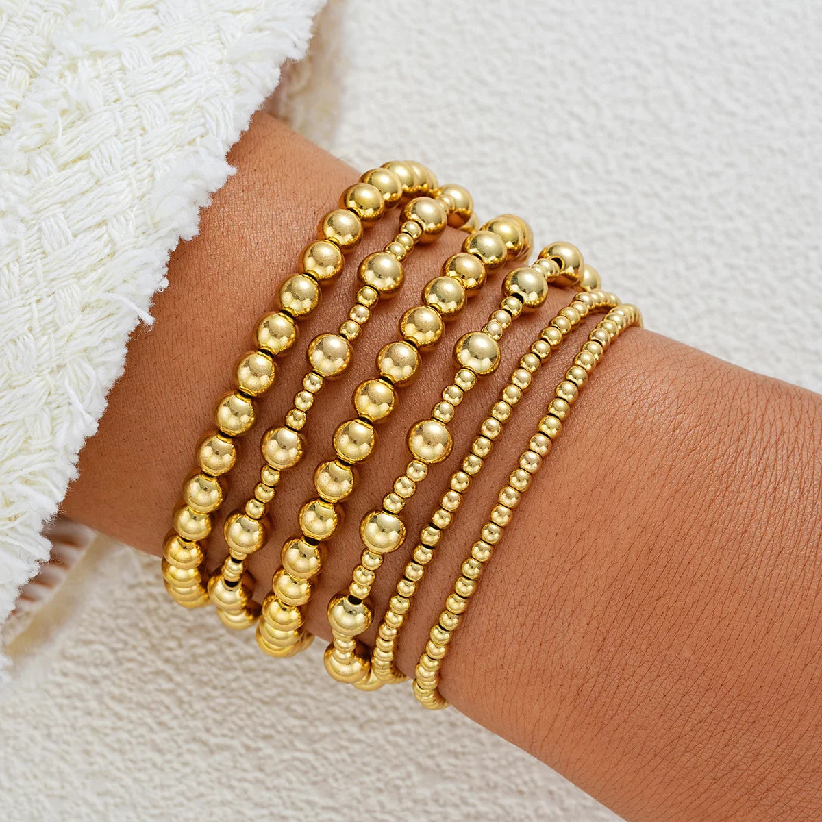 A stack of gold round beaded bracelets worn on a wrist next to a white textured fabric.