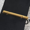 A gold-colored business tie clip with a bar clasp is shown against a dark fabric background.