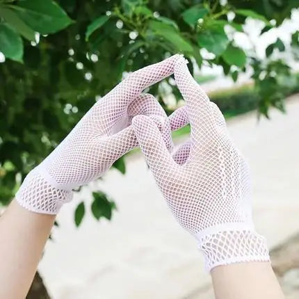 A pair of sheer lace fishnet evening gloves are shown against a blurred green outdoor background.