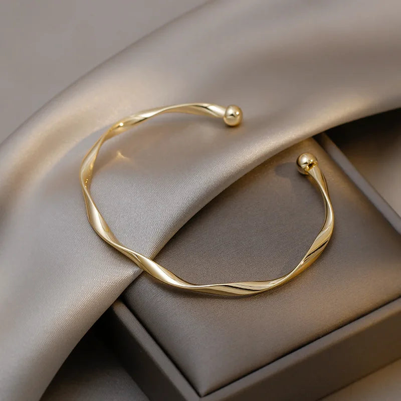 A minimalist gold-tone twisted cuff bangle resting on a neutral gray fabric and box.
