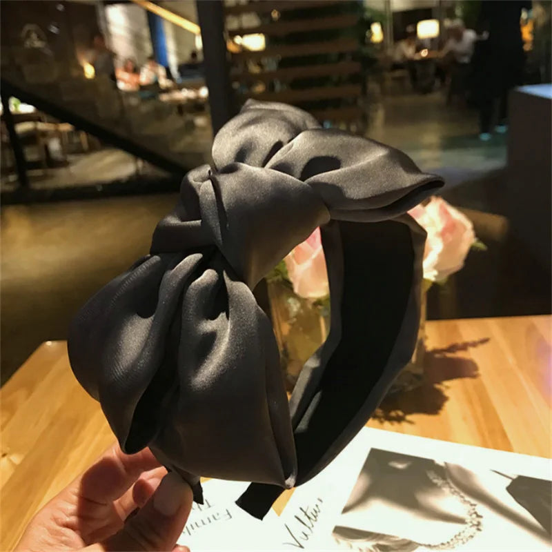 A dark gray chic wide bow headband is held up against a blurred background.