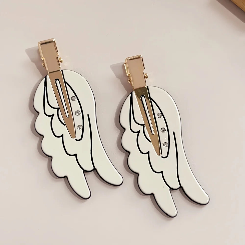 A pair of gold-toned angel wing hair clips with a white, cutout design and small rhinestones.