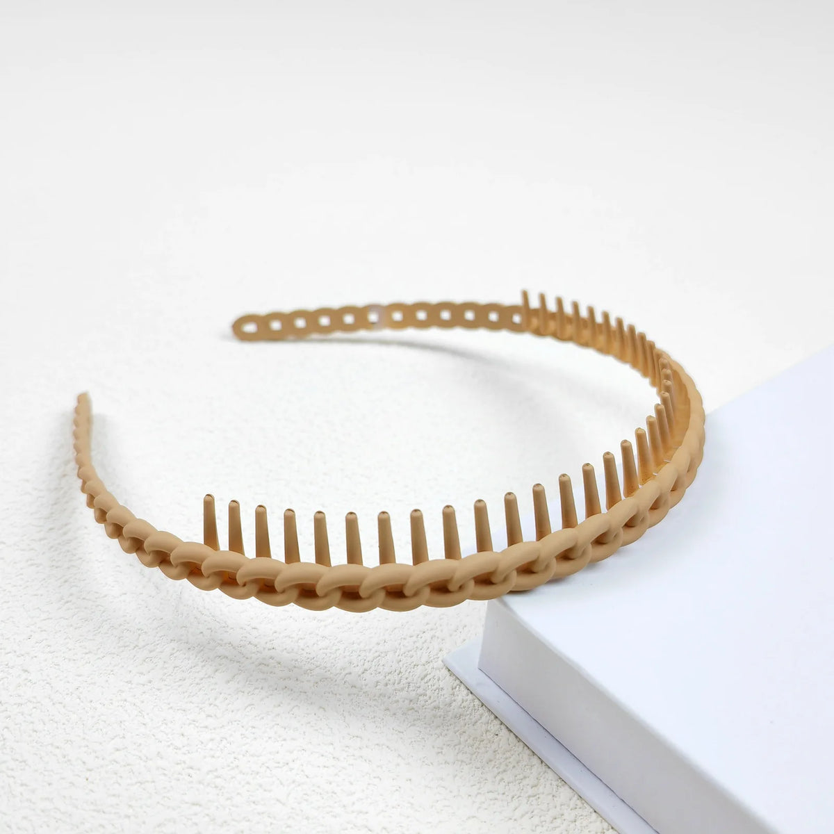 Un bandeau pour cheveux tressé beige avec de petites dents, posé sur une surface blanche.