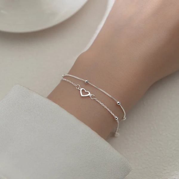 A close-up of a sterling silver double heart charm bracelet worn on a wrist against a light background.
