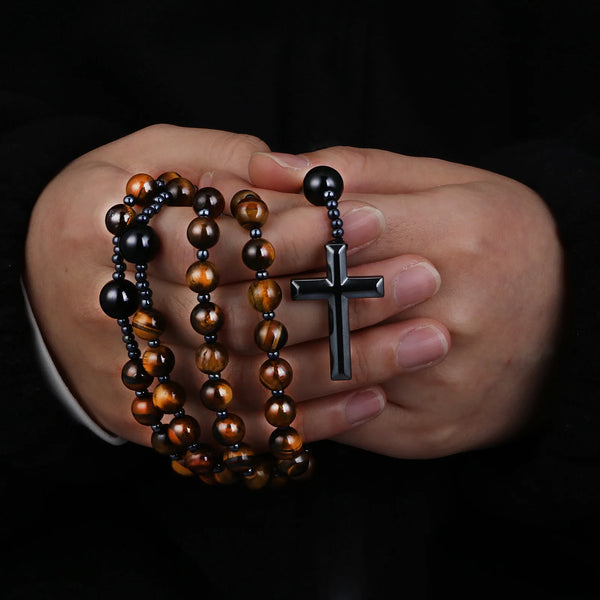 A hand holds a rosary necklace made of tiger eye and black agate beads with a hematite cross pendant.