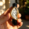 A hand holds a gold-toned quartz watch with an irregular face and a black leather strap.