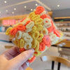 A hand holds a clear plastic package of colorful, fluffy elastic hair bands in shades of yellow, orange, and white.