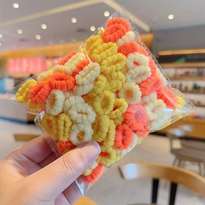 A hand holds a clear plastic package of colorful, fluffy elastic hair bands in shades of yellow, orange, and white.