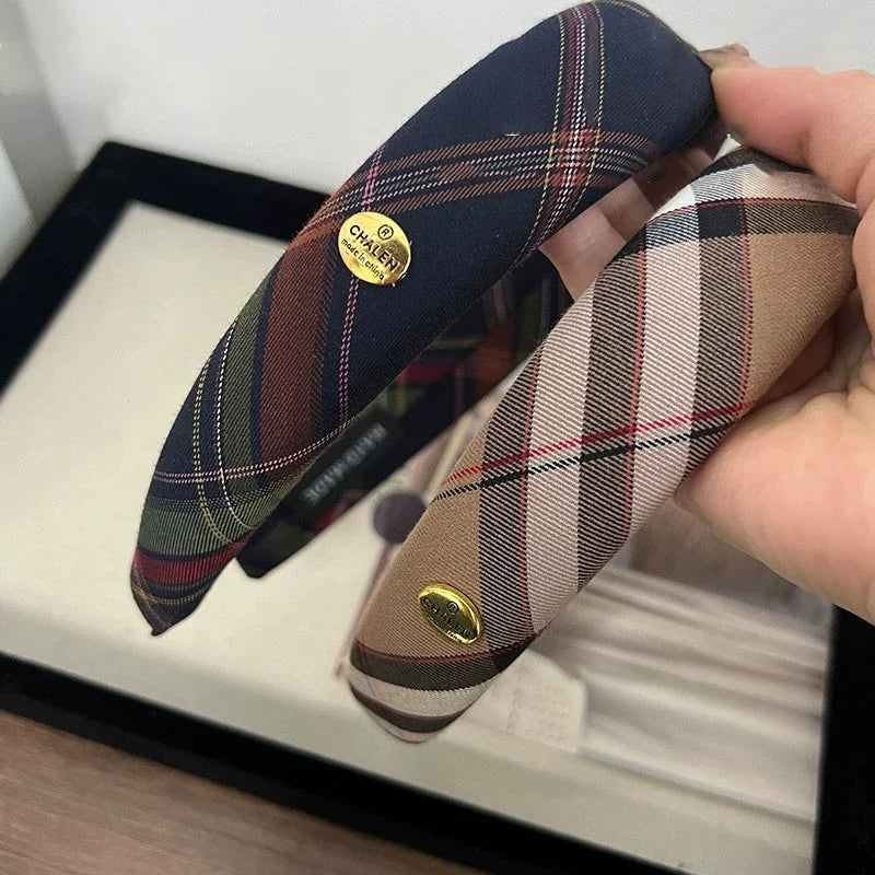 A close-up of two British plaid sponge headbands, one dark blue and another beige, displayed in a black box.