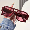 Una mano sostiene unas gafas de sol cuadradas retro marrones con lentes rosadas contra un fondo blanco.