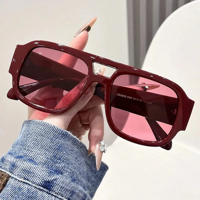 Una mano sostiene unas gafas de sol cuadradas retro marrones con lentes rosadas contra un fondo blanco.
