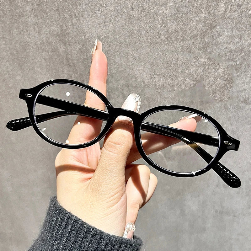A hand holds a pair of black round anti-blue light computer glasses against a gray background.