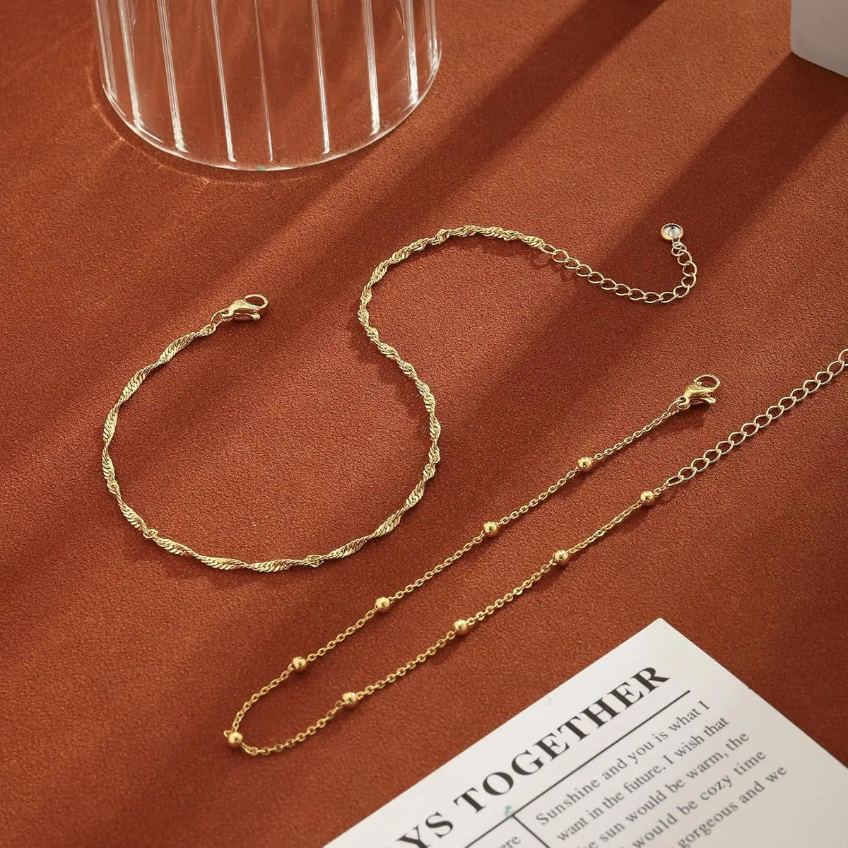 Gold plated beaded anklets displayed on a brown surface next to a glass.
