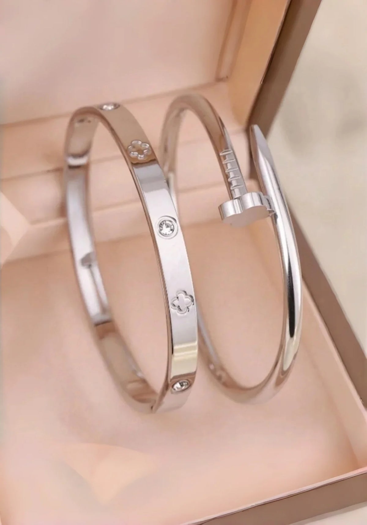 A pair of silver stainless steel bracelets featuring four-leaf clover and nail charms displayed in a light pink box.