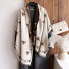 A beige pashmina shawl with a floral print of bees is displayed on a hanger against a wooden background.