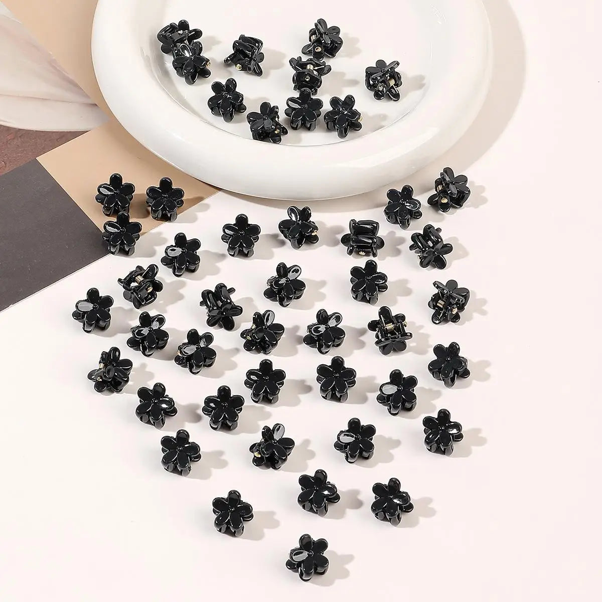 A collection of small black flower-shaped hair clips arranged in a white dish and scattered on a white surface.