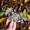 Une main tient une peigne de cheveux de mariée en cristal avec des éléments floraux scintillants délicats.