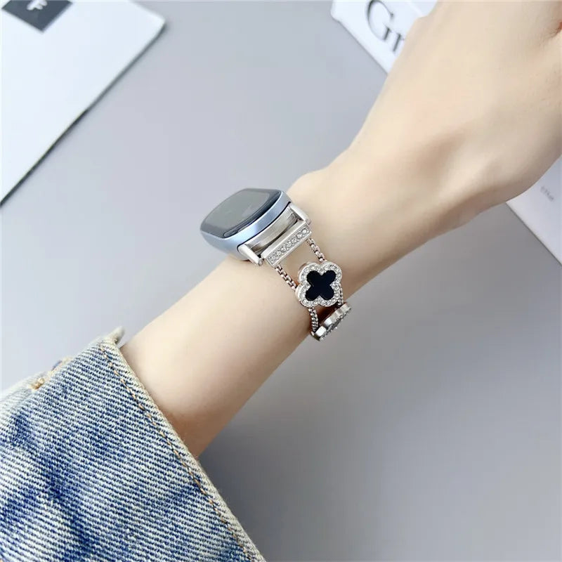 A close-up of a silver watch strap with a diamond four-leaf clover charm on a wrist, next to a denim sleeve.