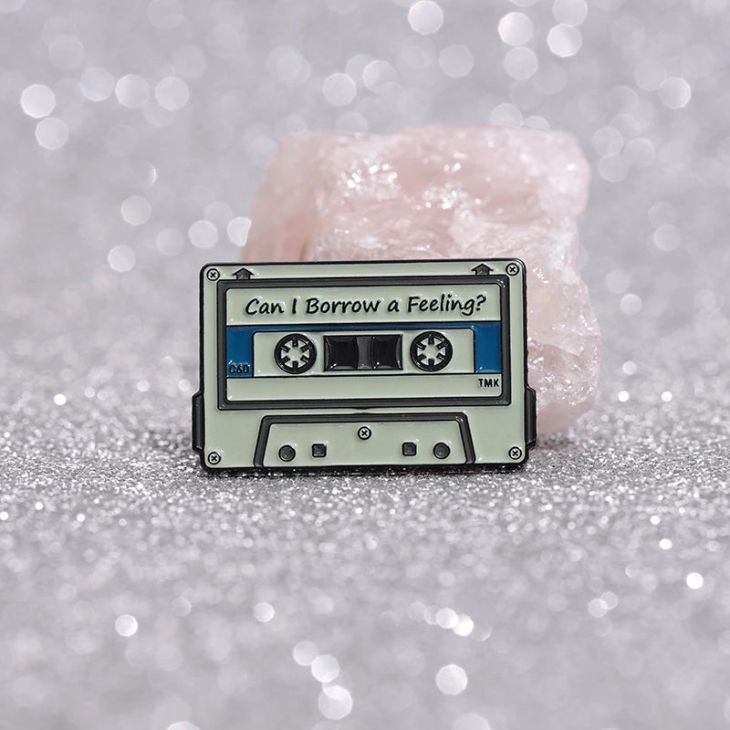 A retro fine line cassette enamel pin brooch with the text Can I Borrow a Feeling on a light pink background.