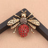 Broche couleur or avec motif abeille, corps texturé et accents de gemme rouge.
