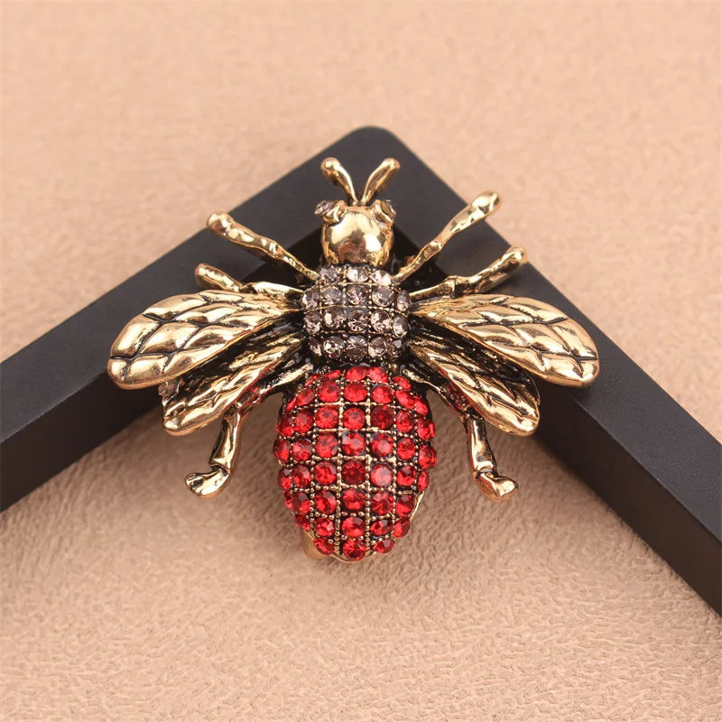 Broche couleur or avec motif abeille, corps texturé et accents de gemme rouge.