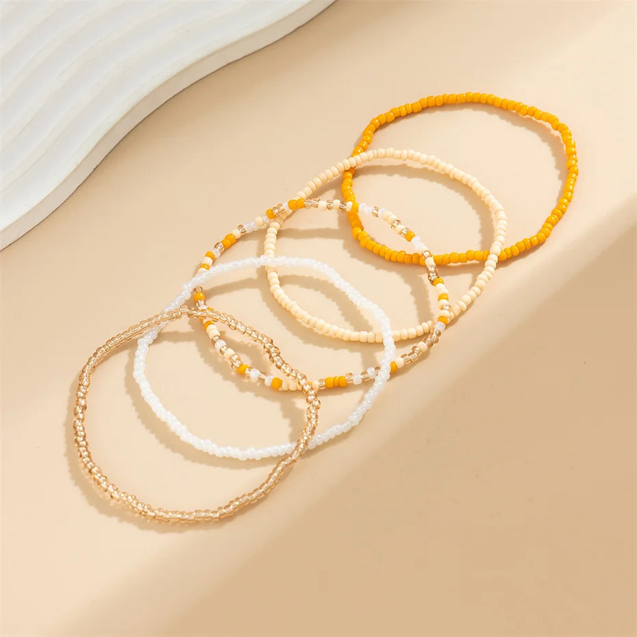 A collection of colorful seed bead anklets and bracelets in shades of yellow, white, and orange laid out on a beige surface.