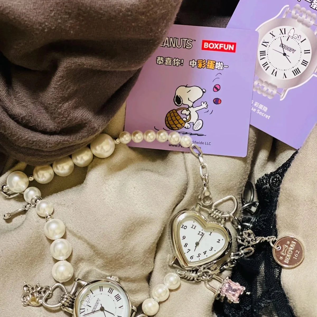 Nahaufnahme einer Perlenkette und einer silbernen Uhr mit Herzanhänger neben einer lila Box mit Snoopy-Grafiken.
