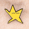 A yellow star shaped enamel pin with the text your did it on a light skin background.