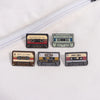 A collection of four retro cassette enamel pins featuring different labels and designs on a white fabric background.
