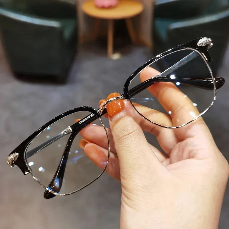 A hand holds a pair of black metal half-frame reading glasses with silver accents against a blurred background.
