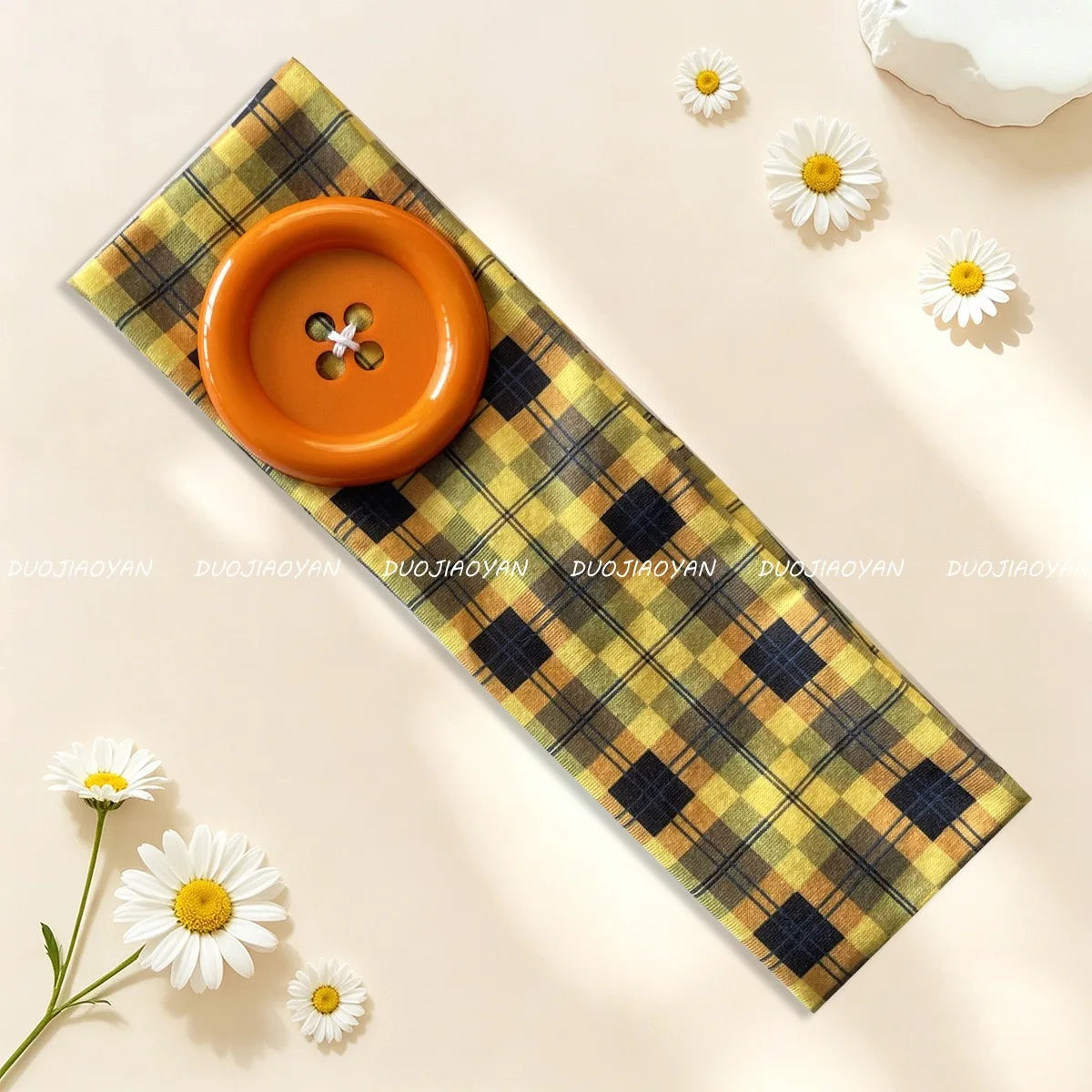 An orange button headband resting on a yellow and black plaid fabric, surrounded by white daisy flowers.