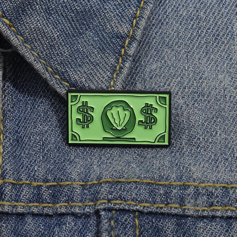 A green enamel pin featuring dollar signs and a leaf symbol pinned onto blue denim fabric.