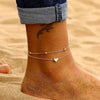 A close-up of a woman's ankle showing an elegant geometric anklet bracelet on her skin against a sandy background.
