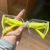 A hand holds a pair of bright yellow fashion glasses with clear frames against a blurred background.