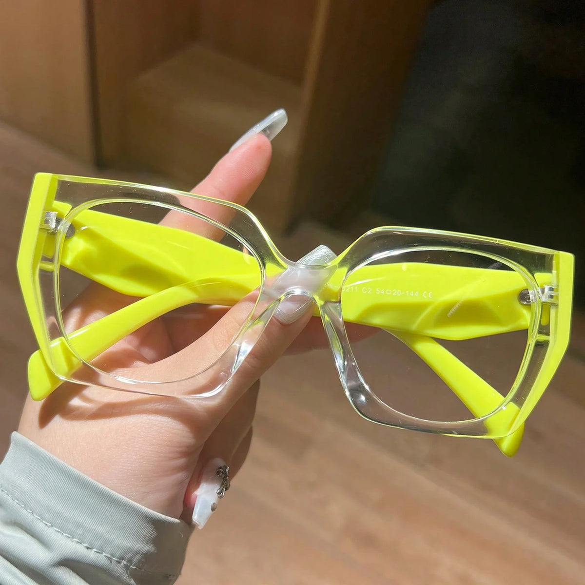 A hand holds a pair of bright yellow fashion glasses with clear frames against a blurred background.