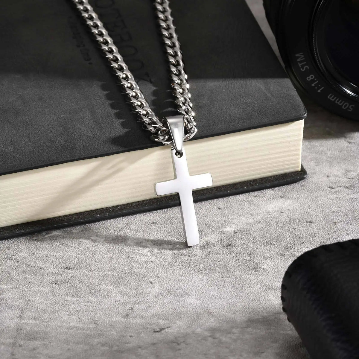 A silver stainless steel Cuban link necklace with a cross pendant displayed on a dark book.