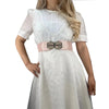 White lace-trimmed dress with sheer sleeves and a light pink elastic belt featuring a brass buckle