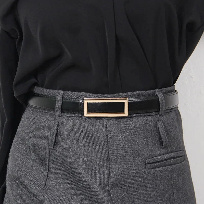 A close-up of a black blouse and gray trousers featuring a black leather belt with a gold buckle.