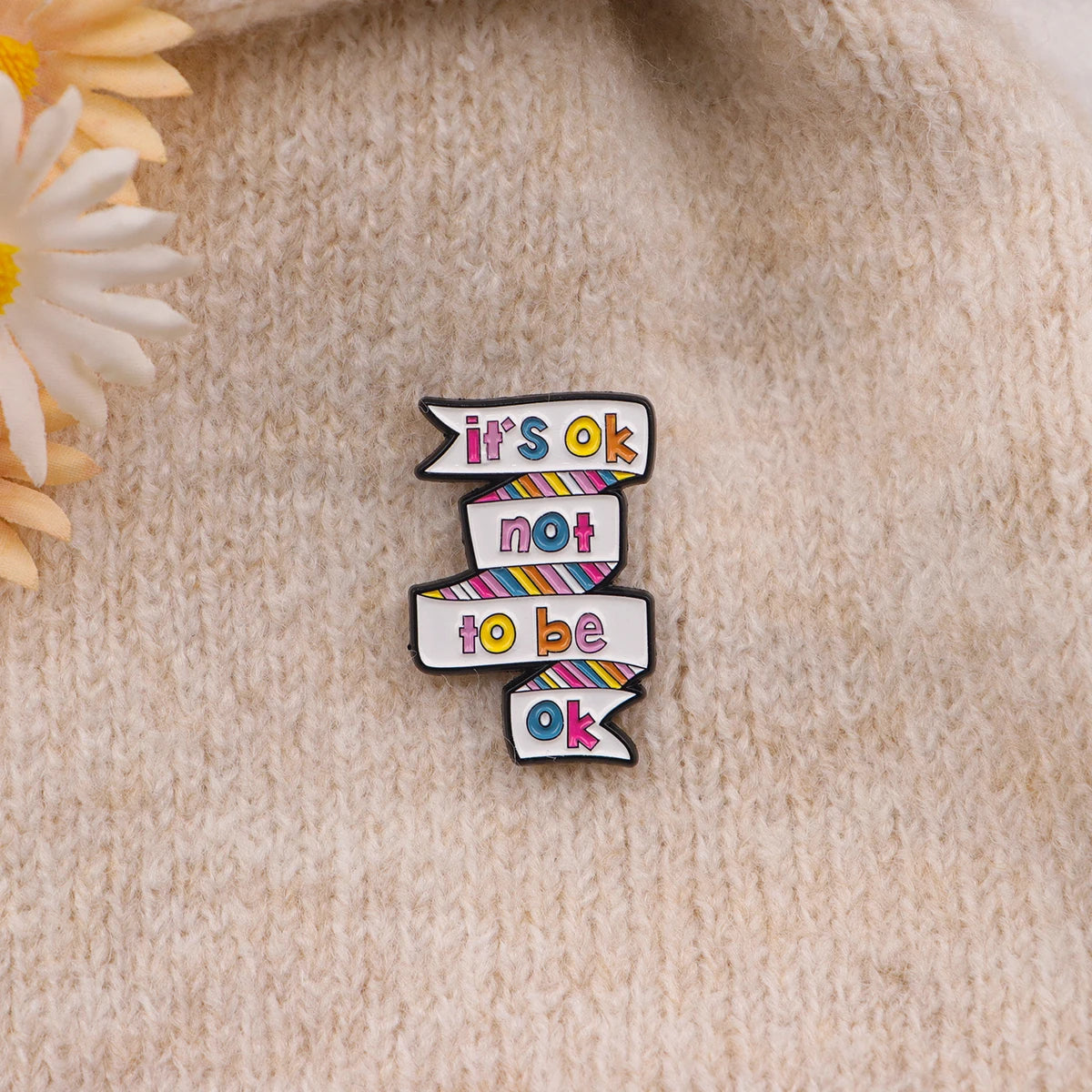 A black enamel pin featuring the text it's ok not to be ok in multicolored ribbon style.