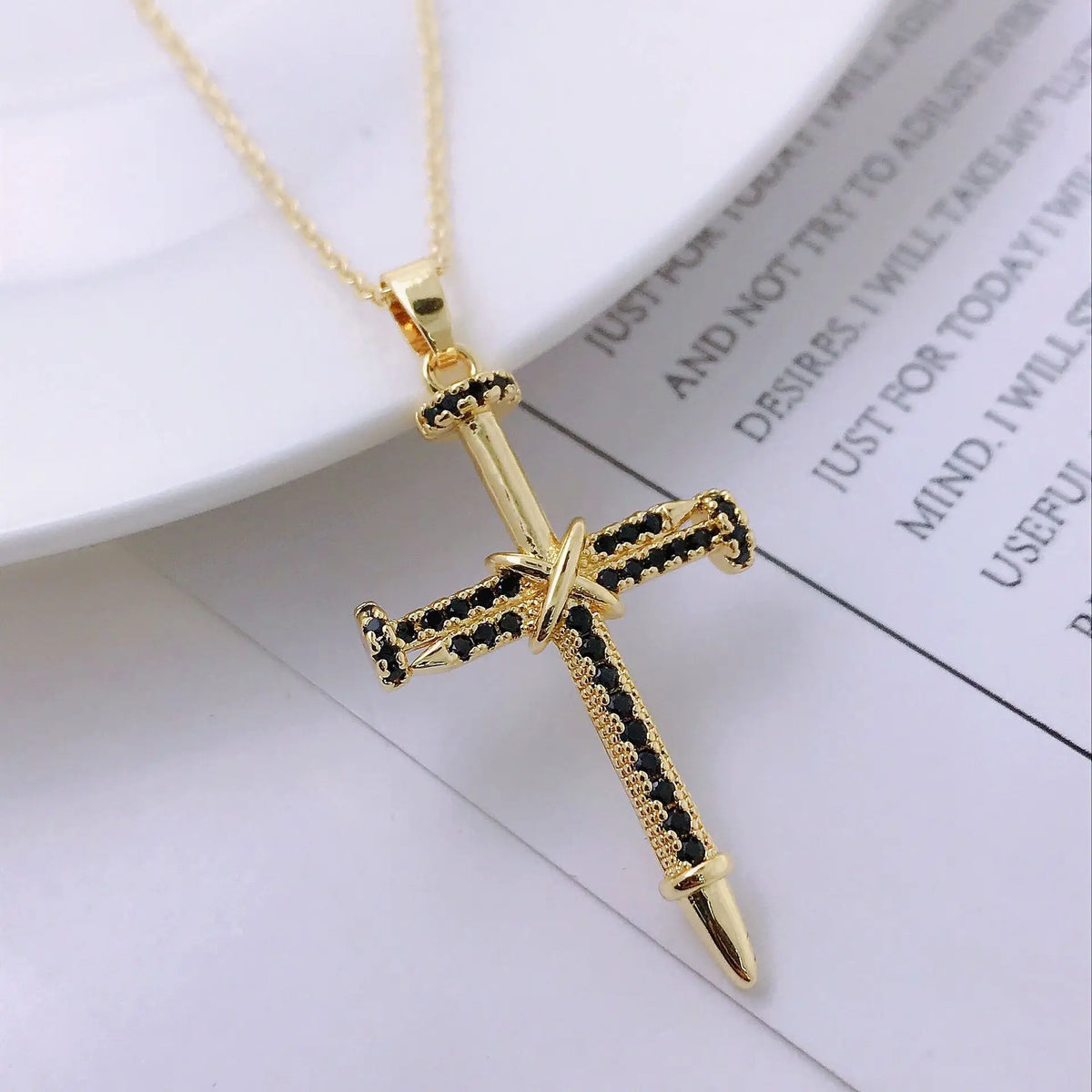 A dainty gold cross charm necklace with black and gold detailing is displayed on a white background.
