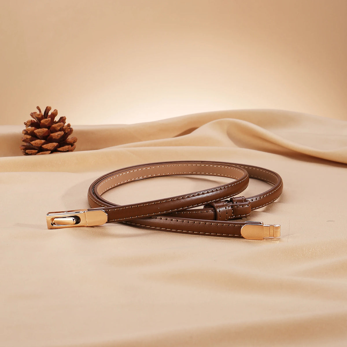 A close-up of a brown skinny leather belt with an adjustable gold buckle lying on a beige fabric.