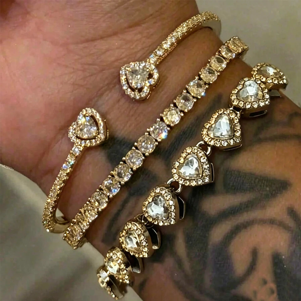 A set of gold-toned geometric rhinestone heart stacking bracelets worn on a hand.