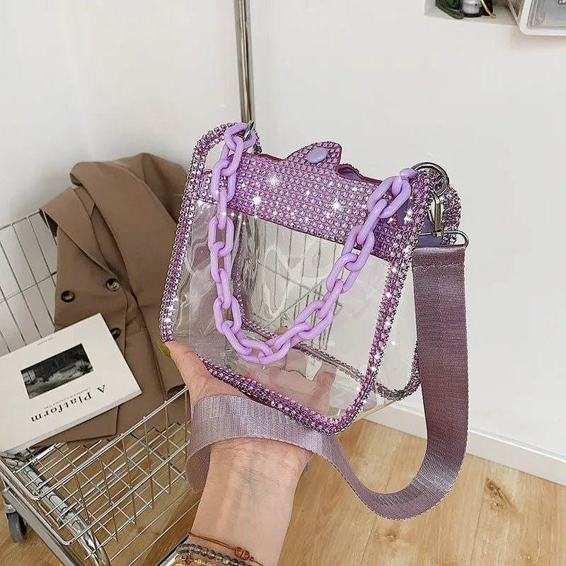Bolso cruz transparente de PVC morado con detalles de cadena plateada, junto a un abrigo y una canasta de alambre.