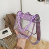 Bolso cruz transparente de PVC morado con detalles de cadena plateada, junto a un abrigo y una canasta de alambre.