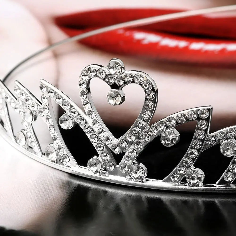 A silver crystal hair crown for women with a heart design resting on a dark surface near red lips.